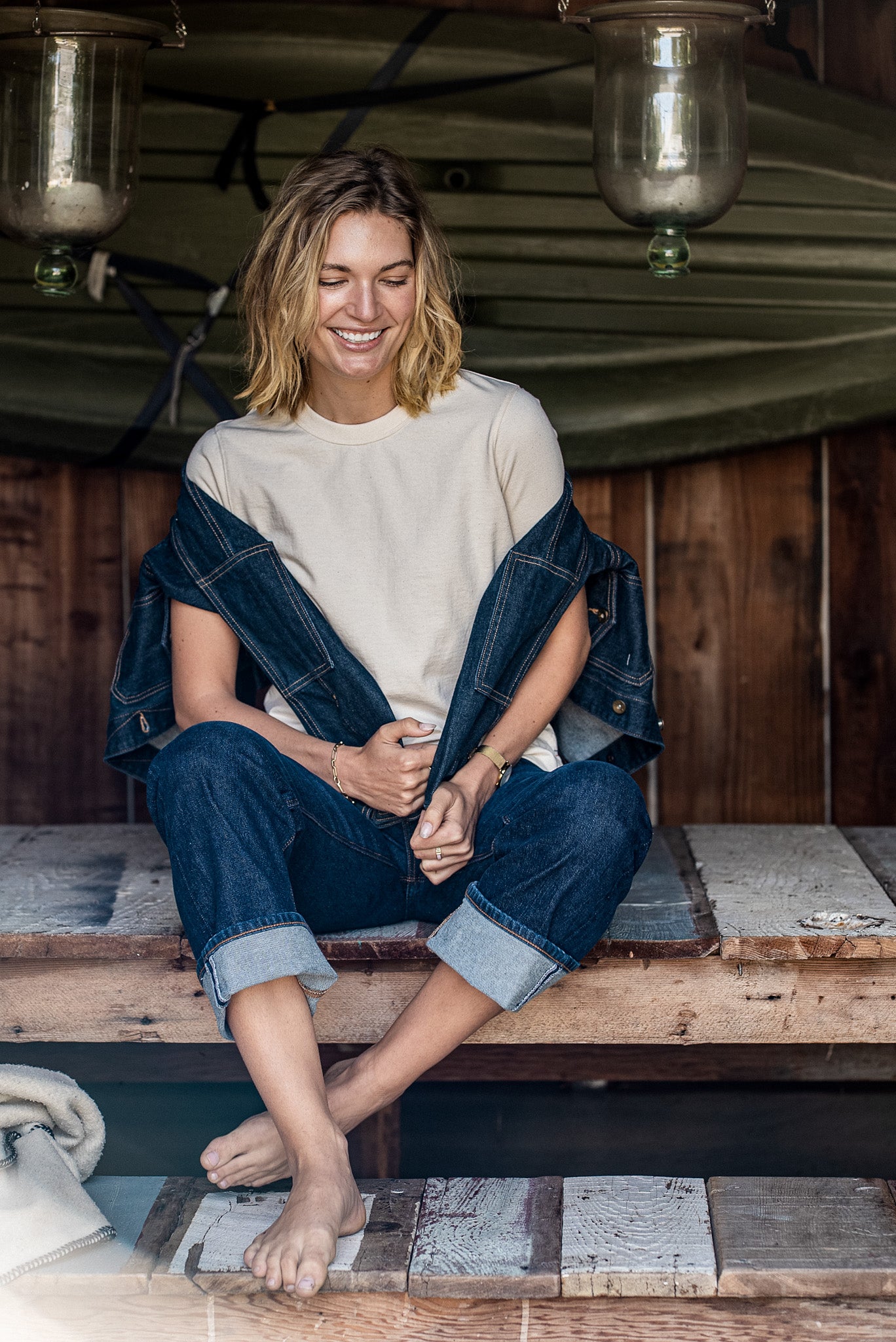 Smiling woman in natural Reyes boxy tee, raw denim jacket and cuffed jeans sitting on a wooden porch.