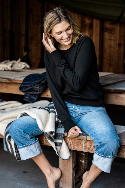 Woman in a black rugby jersey pullover and cuffed selvedge denim jeans sitting on a wooden bench.