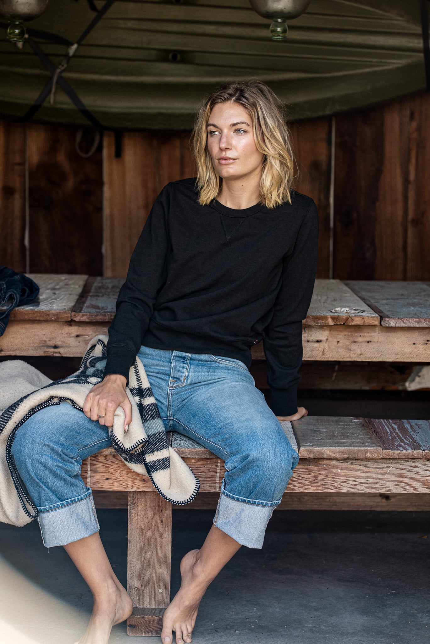 Model in black Stinson rugby jersey pullover and cuffed selvedge denim jeans, seated on a wooden bench.