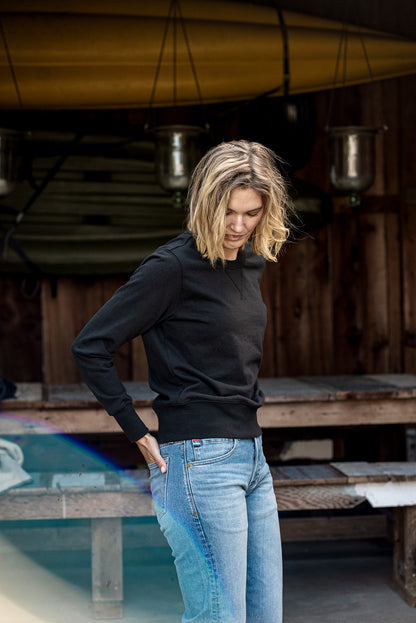 Woman wearing the black Stinson rugby jersey pullover with light-wash jeans in a boathouse.