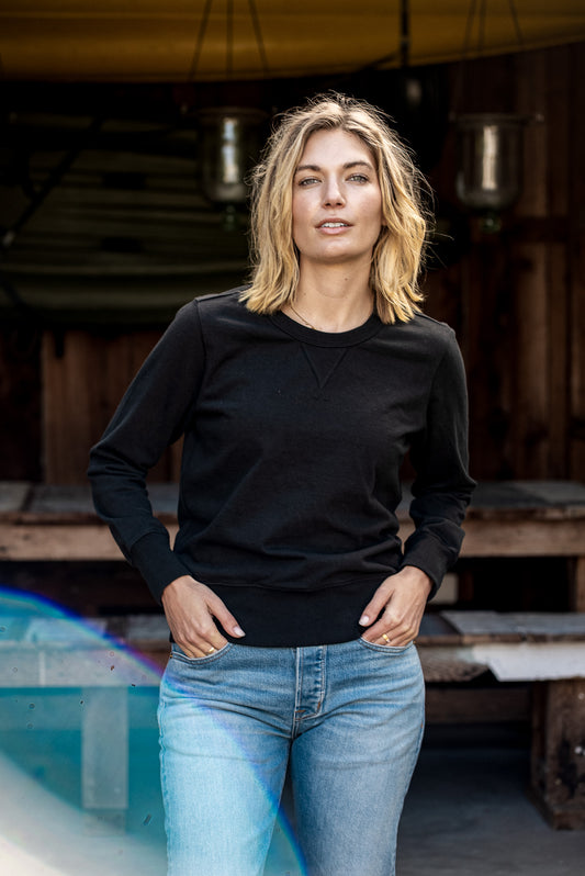 Woman in a basalt black rugby jersey pullover and light-wash jeans standing outside a boathouse.