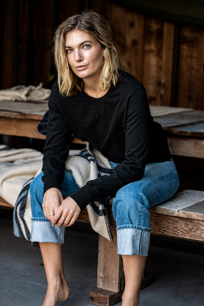 Model in black Stinson rugby jersey pullover and cuffed selvedge denim jeans sitting on a wooden bench.