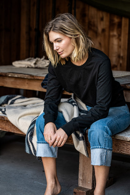 Woman in a black rugby jersey pullover and cuffed blue jeans seated on a wooden bench.