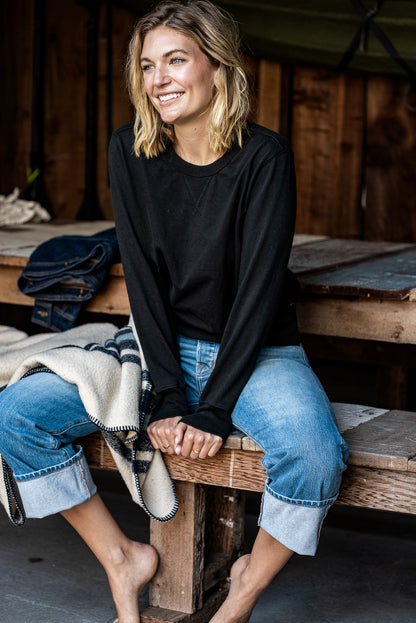 Smiling person in a black Stinson rugby jersey pullover and cuffed selvedge denim jeans, seated on a wooden bench.