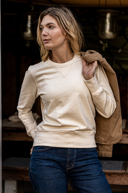 Woman wearing a natural cream Stinson rugby jersey pullover with dark denim jeans, holding a tan jacket.