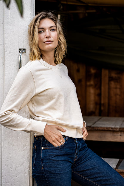 Woman in a natural cream Stinson rugby jersey pullover and dark denim jeans leaning against a white barn door.