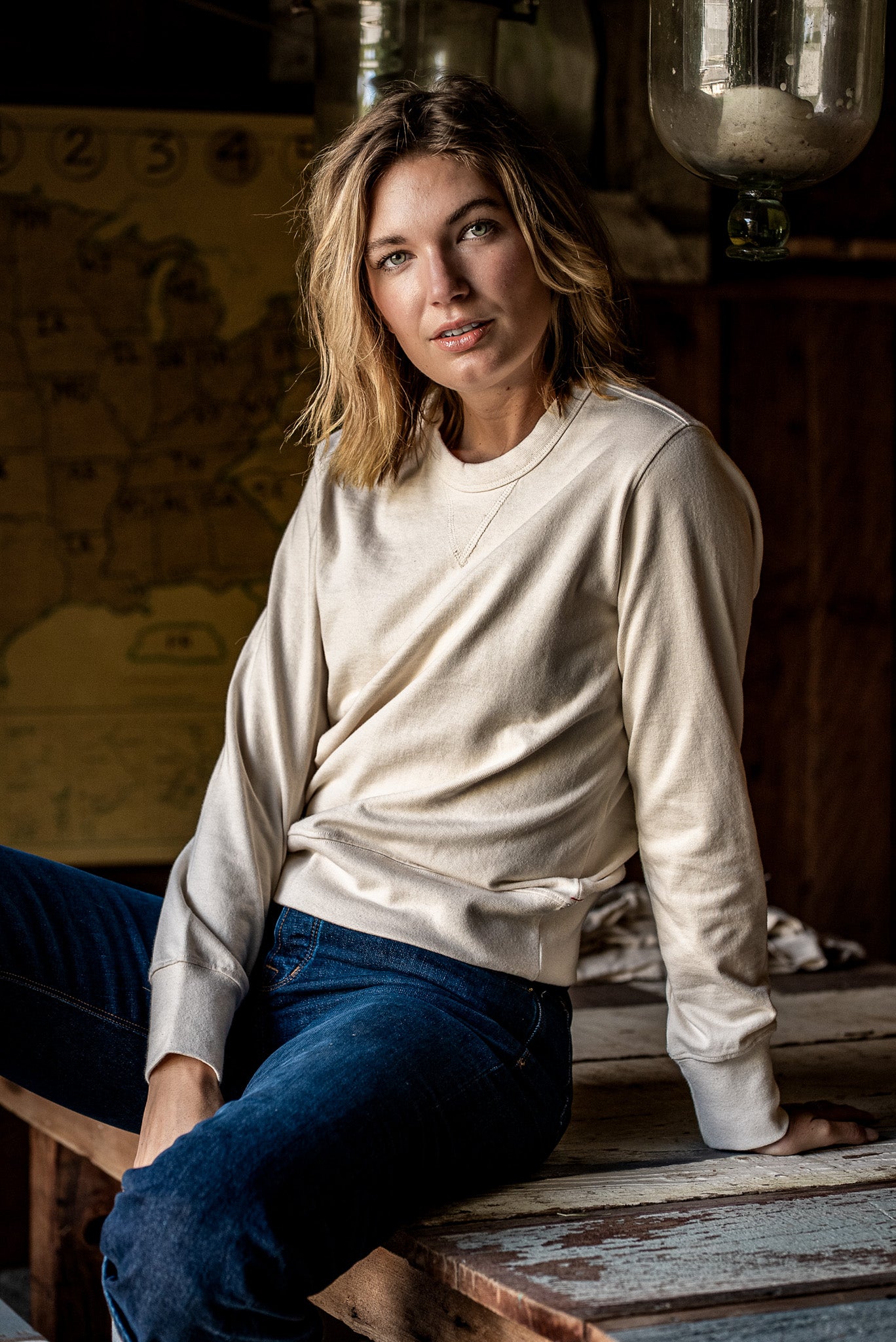 Woman in a natural cream Stinson rugby jersey pullover and dark denim jeans, seated in a rustic workshop.