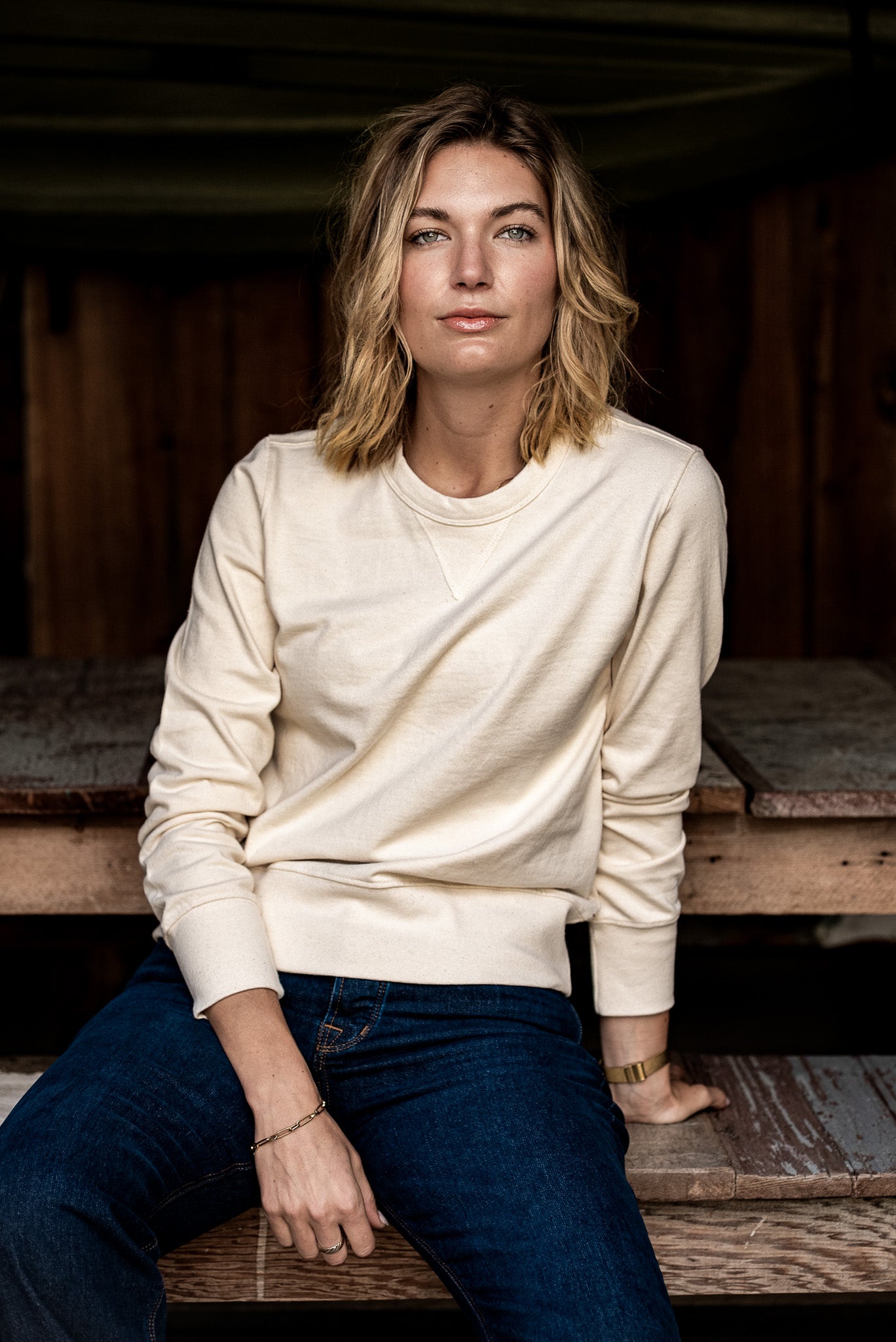 Woman in a natural cream Stinson rugby jersey pullover and dark denim jeans seated on a wooden bench.