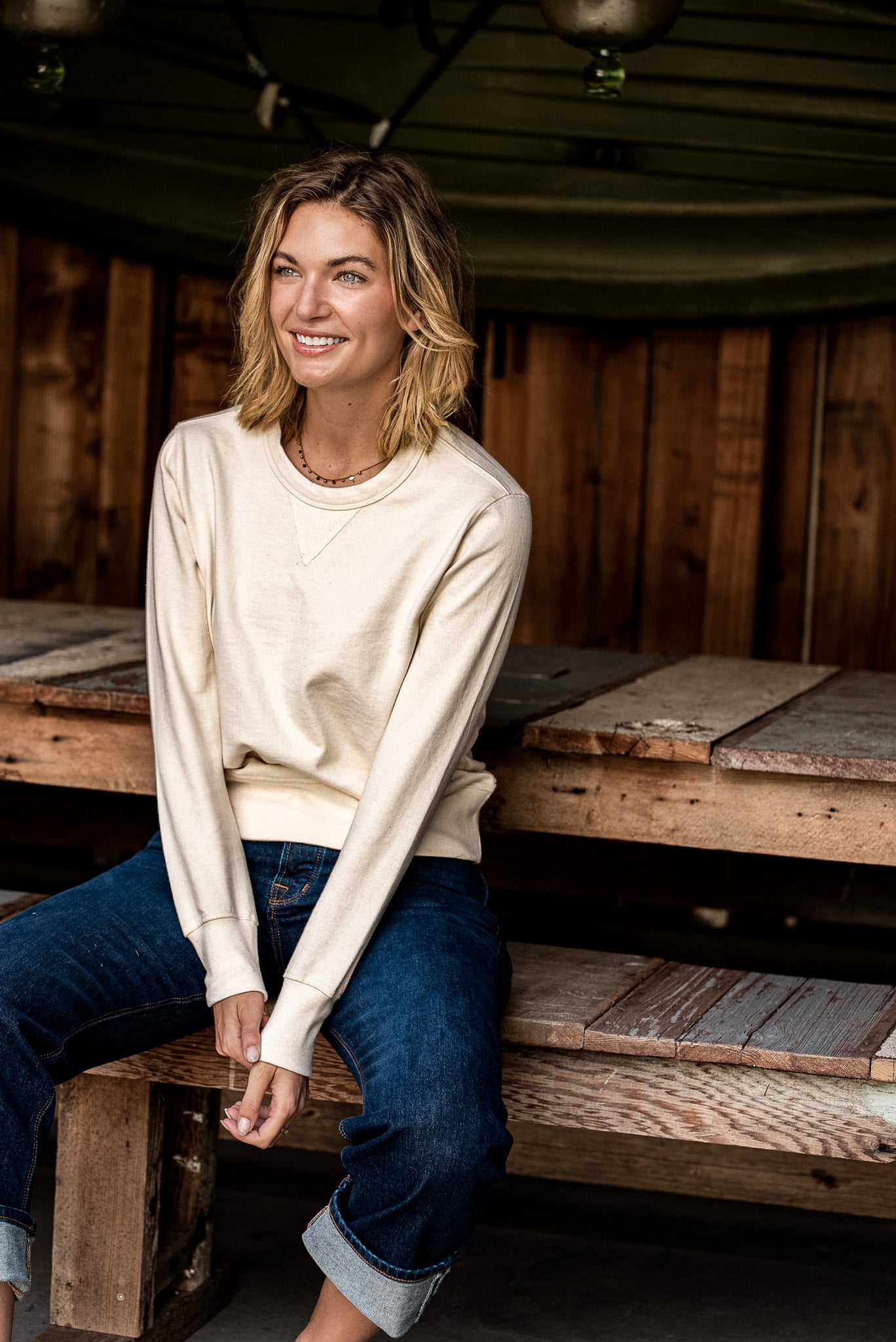 Woman in a natural cream Stinson rugby jersey pullover and cuffed selvedge denim jeans, seated on a wooden bench.