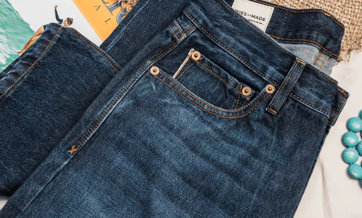 Sidney Fit Jean in Ocean Washed color, featuring a high-rise straight leg style, displayed on a textured surface with a woven mat, turquoise beads, & a surf magazine in the background