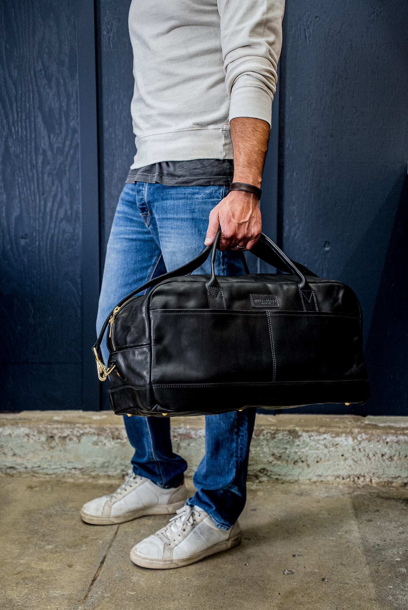 Person carrying a black Horween leather weekender bag with brass zipper by the handles.