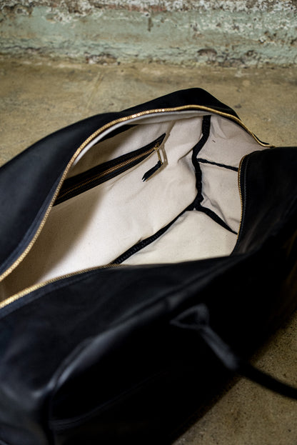 Interior of black leather weekender bag showing natural cotton canvas lining and brass zip pocket.