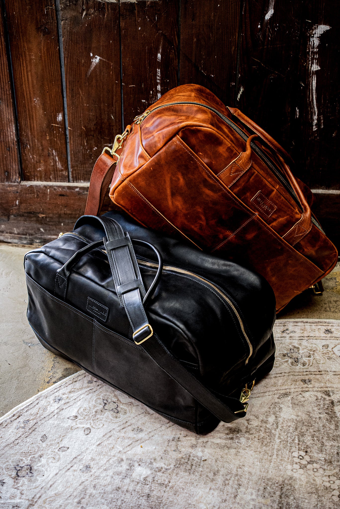 Black Horween leather weekender bag with brass zipper, resting beside a tan leather duffel.