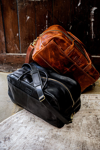 Black Horween leather weekender bag with brass zipper, resting beside a tan leather duffel.