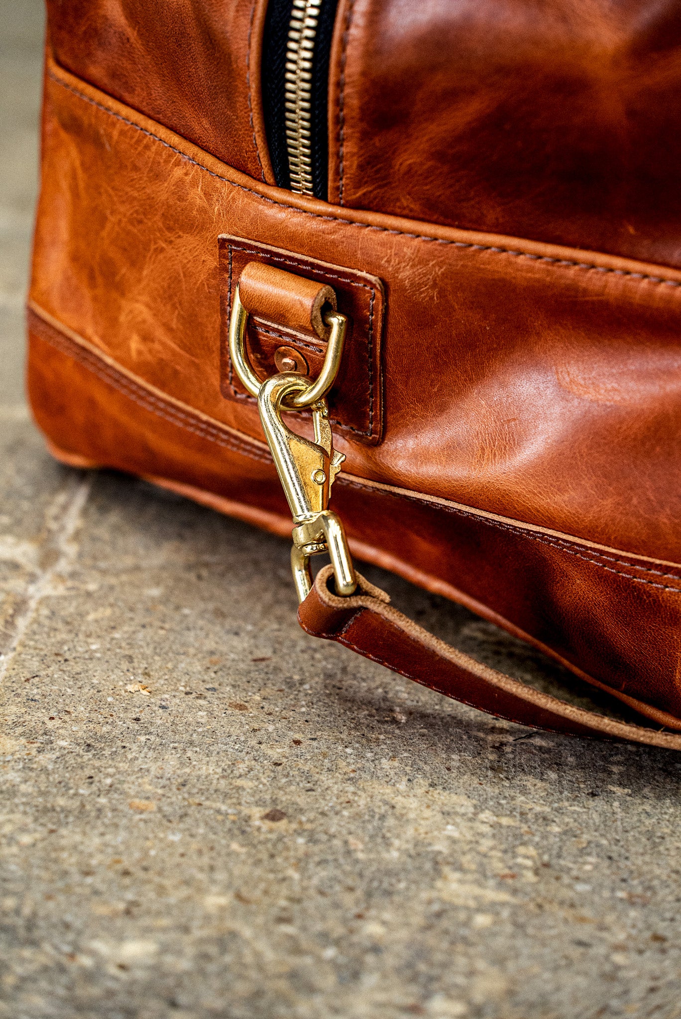 Brass swivel hook clipping the shoulder strap onto a tan leather weekender bag.