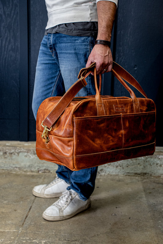 Tan Horween leather weekender bag with brass hardware, carried by a person in denim.