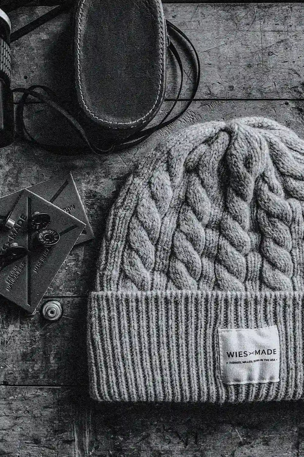 Natural cable-knit wool beanie with ribbed cuff and WiesMade label, on weathered wood beside leather goods.
