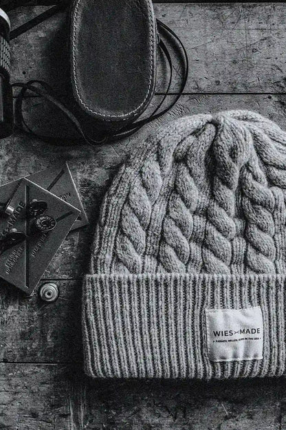 Natural cable-knit wool beanie with ribbed cuff and WiesMade label, on weathered wood beside leather goods.
