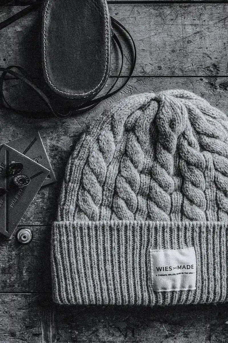 Heather gray cable-knit wool beanie with WiesMade label, styled on weathered wood beside leather boots.
