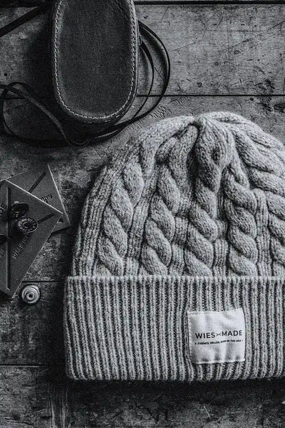 Gray cable-knit wool beanie with WiesMade label, resting on weathered wood beside leather boots.