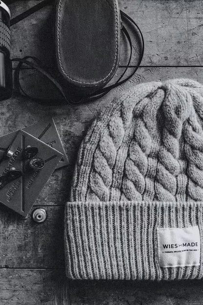 Cable-knit wool beanie with folded cuff and WiesMade label on a wooden surface beside leather goods.