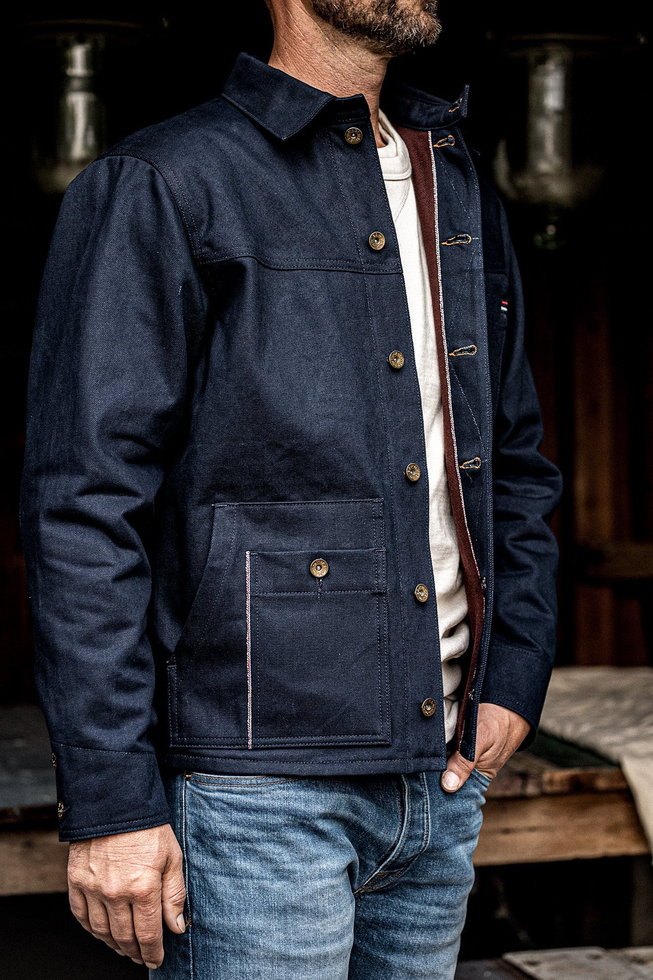 Man wearing a WiesMade raw indigo denim chore coat with brass buttons over a white tee and faded jeans.