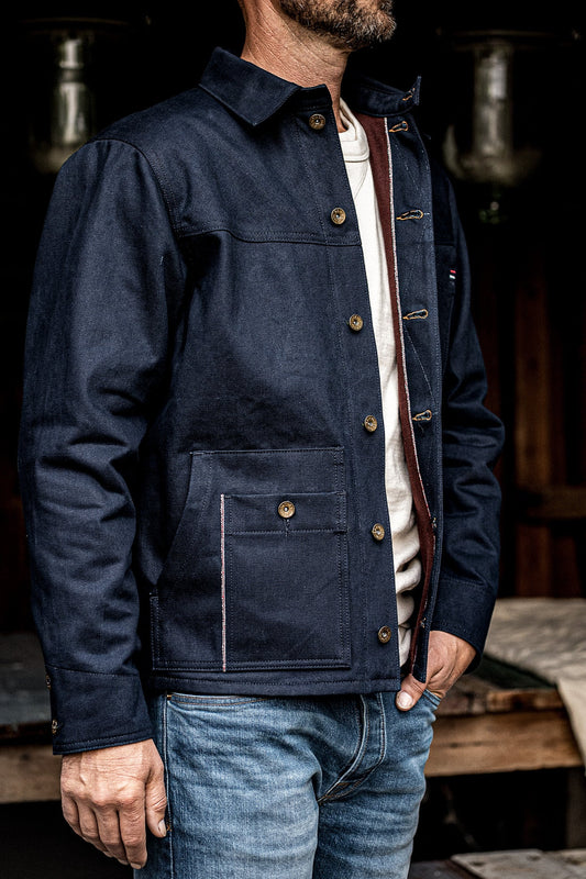 Man wearing a WiesMade raw indigo denim chore coat with brass buttons over a white tee and faded jeans.