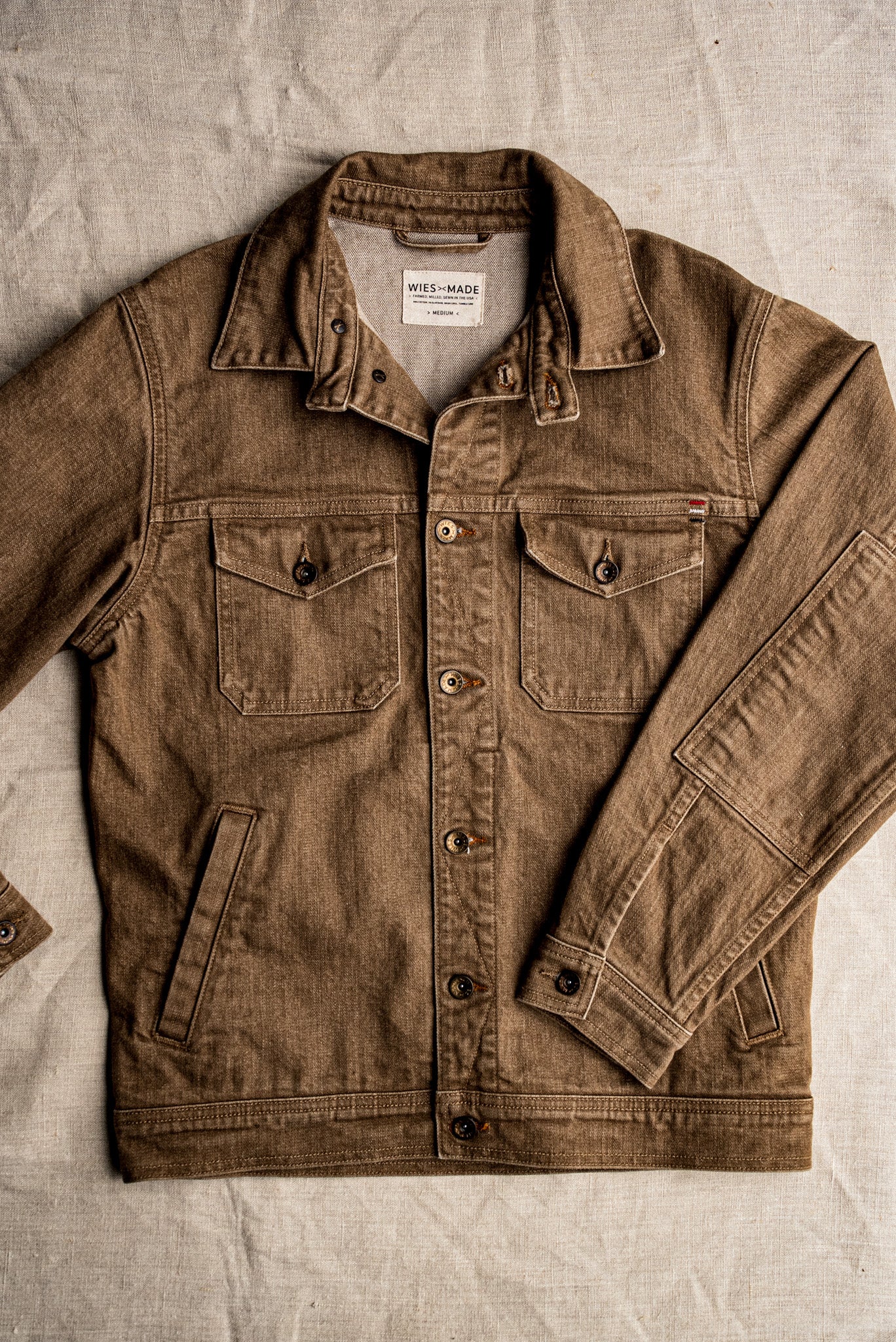 Wheat-colored broken twill denim jacket laid flat on linen, showing button front, chest flap pockets, and brass hardware.