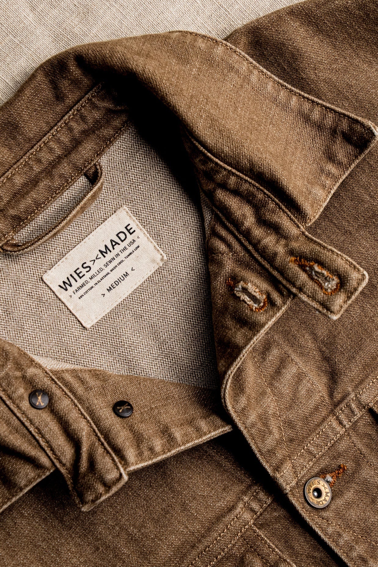 Close-up of wheat-colored broken twill denim jacket showing interior WiesMade label, tobacco buttonholes, and brass hardware.