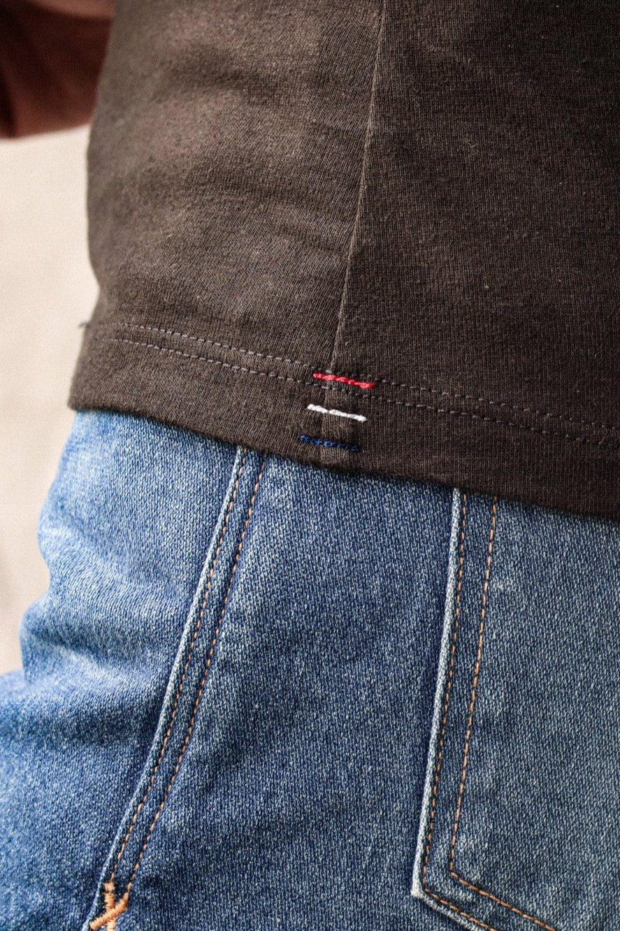 Close-up of basalt brown cotton tank hem with red, white, and blue signature bartacks over blue jeans.