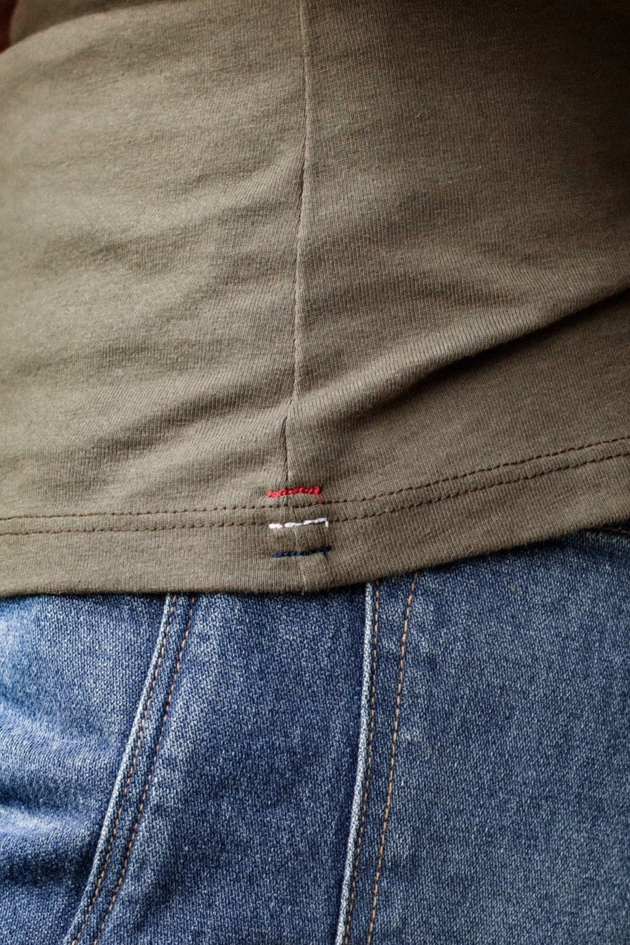 Close-up of olive cotton Bolinas tank hem with red, white, and blue signature triple bartack stitching over blue denim jeans.