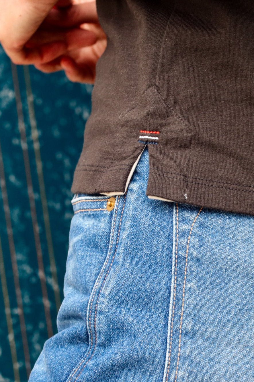 Close-up of the Reyes tank's side vent hem with signature triple bartack stitching, worn over blue denim jeans.