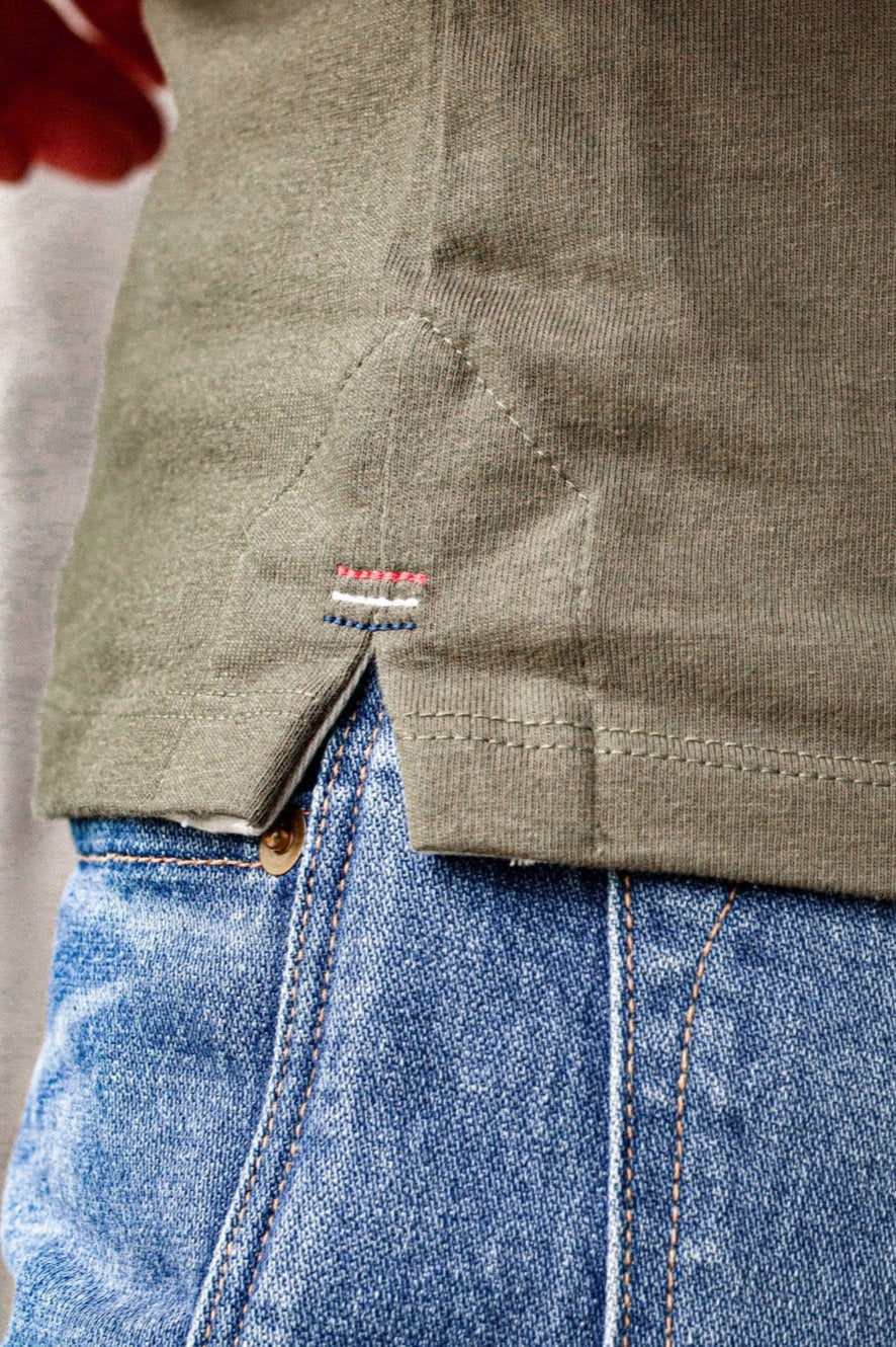 Close-up of the olive cotton Reyes tank hem showing signature red, white, and blue triple bartack stitching over blue denim.