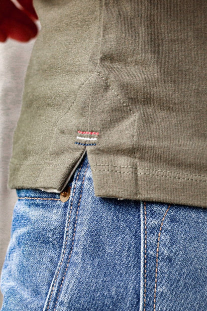 Close-up of the olive cotton Reyes tank hem showing signature red, white, and blue triple bartack stitching over blue denim.