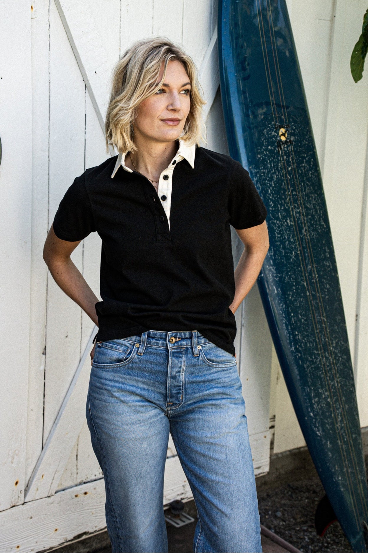 Woman in a black short-sleeve rugby jersey with white collar and jeans, standing beside a surfboard.