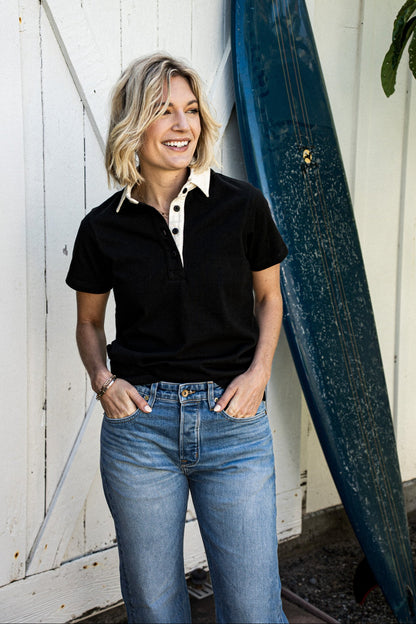 Woman in a black short-sleeve rugby jersey with cream collar and jeans, leaning against a white barn door beside a surfboard.