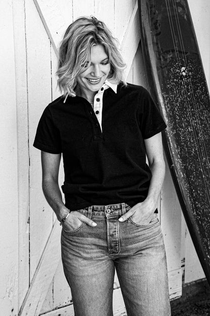 Woman in a black short-sleeve rugby jersey and jeans, standing beside a surfboard against a white barn door.