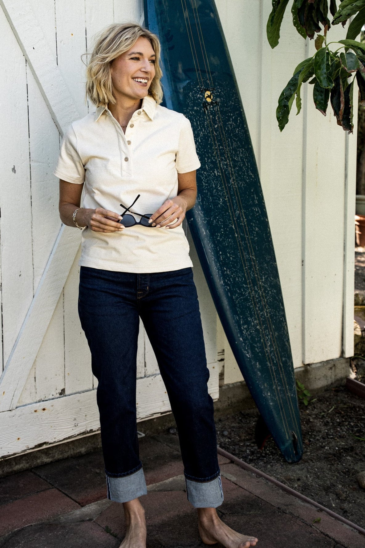 Woman in a natural cotton women's rugby jersey, holding sunglasses beside a leaning surfboard.
