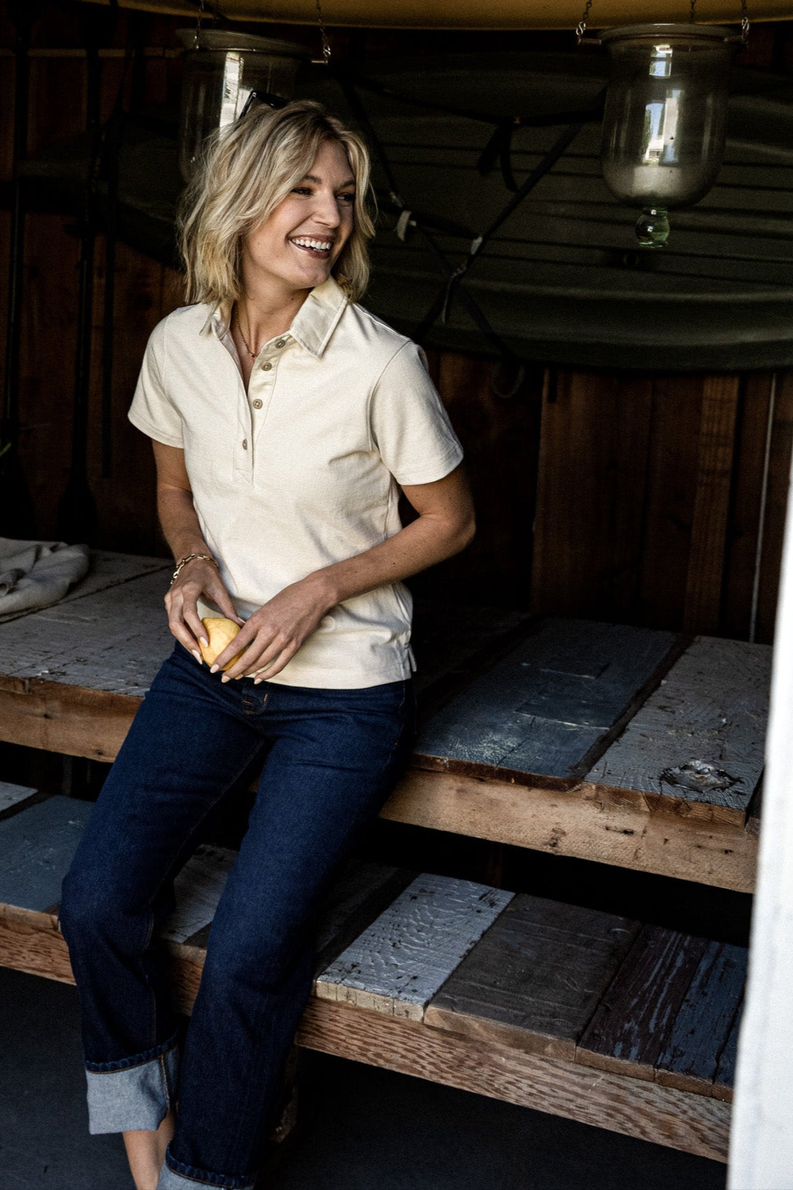 Woman in a natural cream short-sleeve rugby polo and cuffed dark denim jeans, leaning against wooden steps.