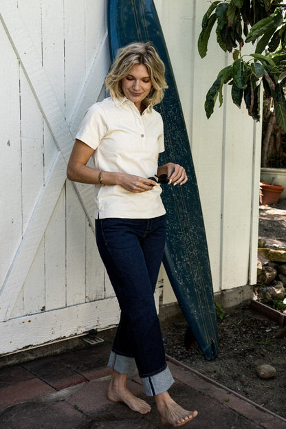 Woman in a natural rugby jersey and cuffed denim jeans leaning by a vintage surfboard.