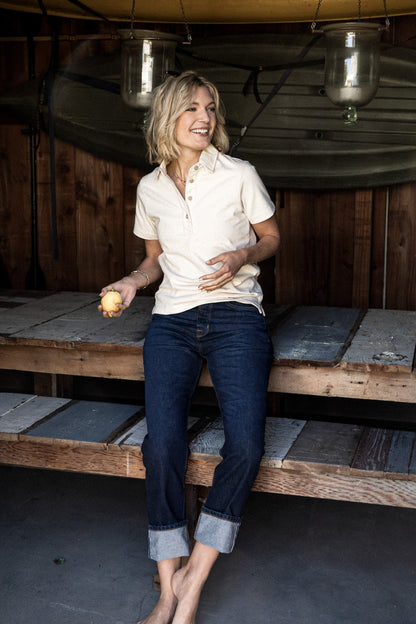 Woman in a natural cream short-sleeve rugby jersey and cuffed dark denim jeans, seated on a wooden bench.