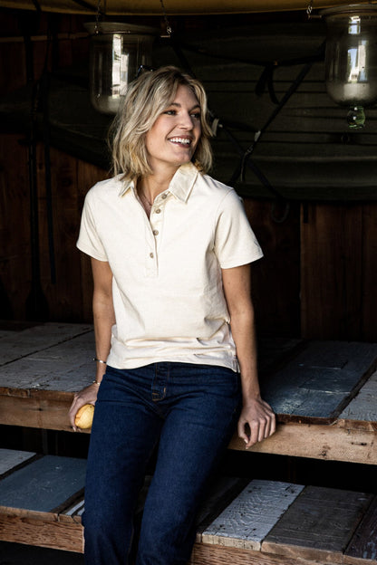 Smiling woman in a natural short-sleeve cotton rugby polo and dark jeans, seated on wooden benches.