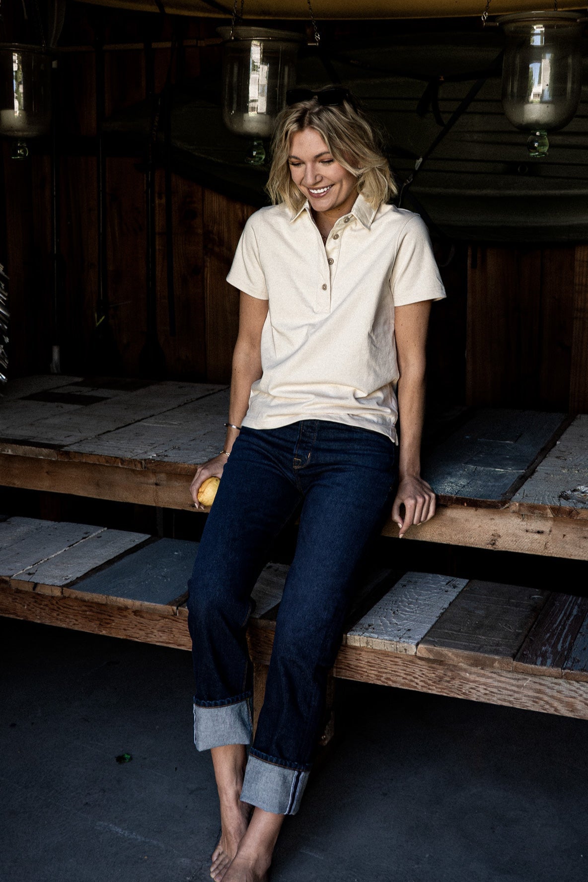 Woman in a natural cotton short-sleeve rugby jersey and cuffed dark denim jeans, seated on a wooden platform.