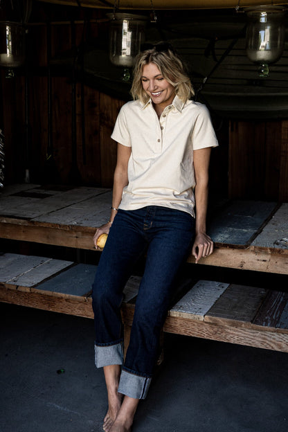 Woman in a natural cotton short-sleeve rugby jersey and cuffed dark denim jeans, seated on a wooden platform.