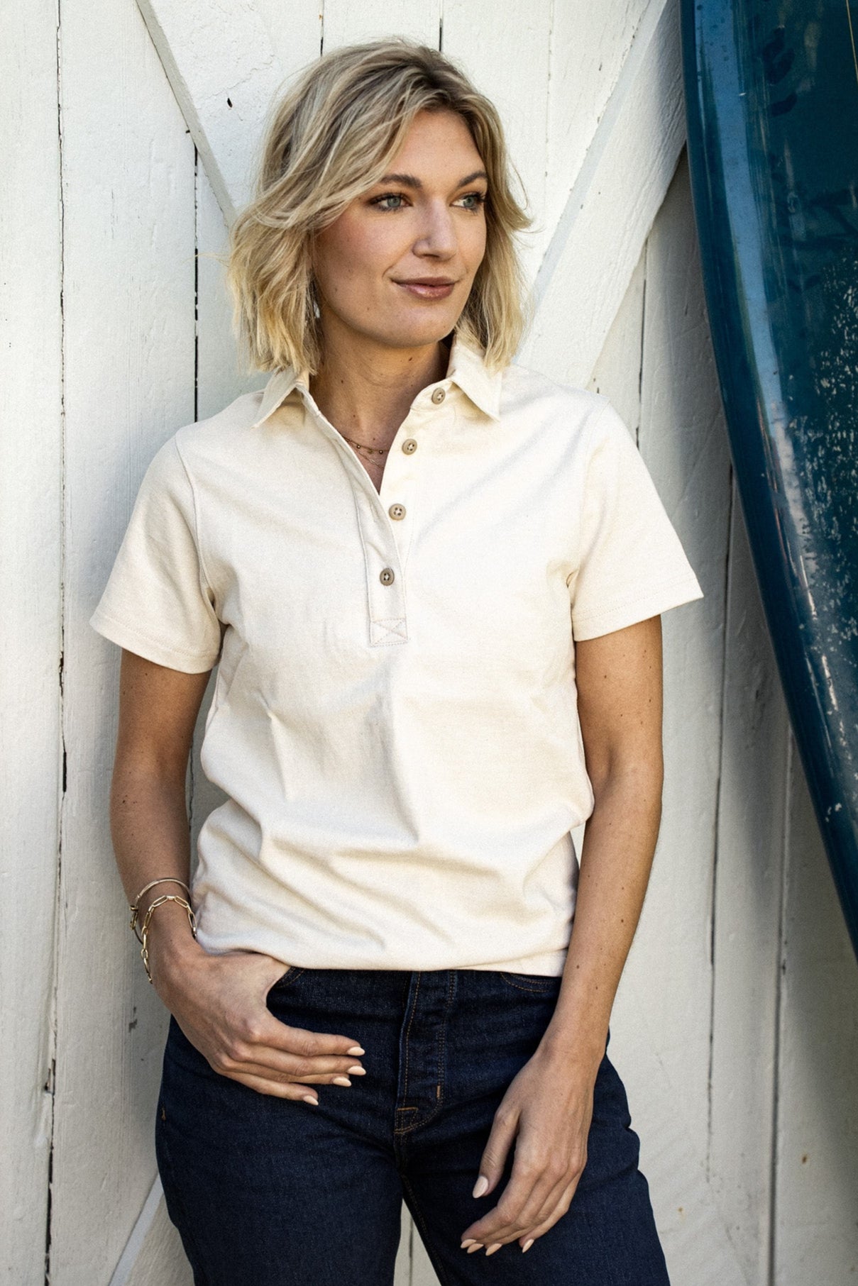 Woman in a natural cream short-sleeve rugby jersey with button placket, leaning against a white wall beside a surfboard.