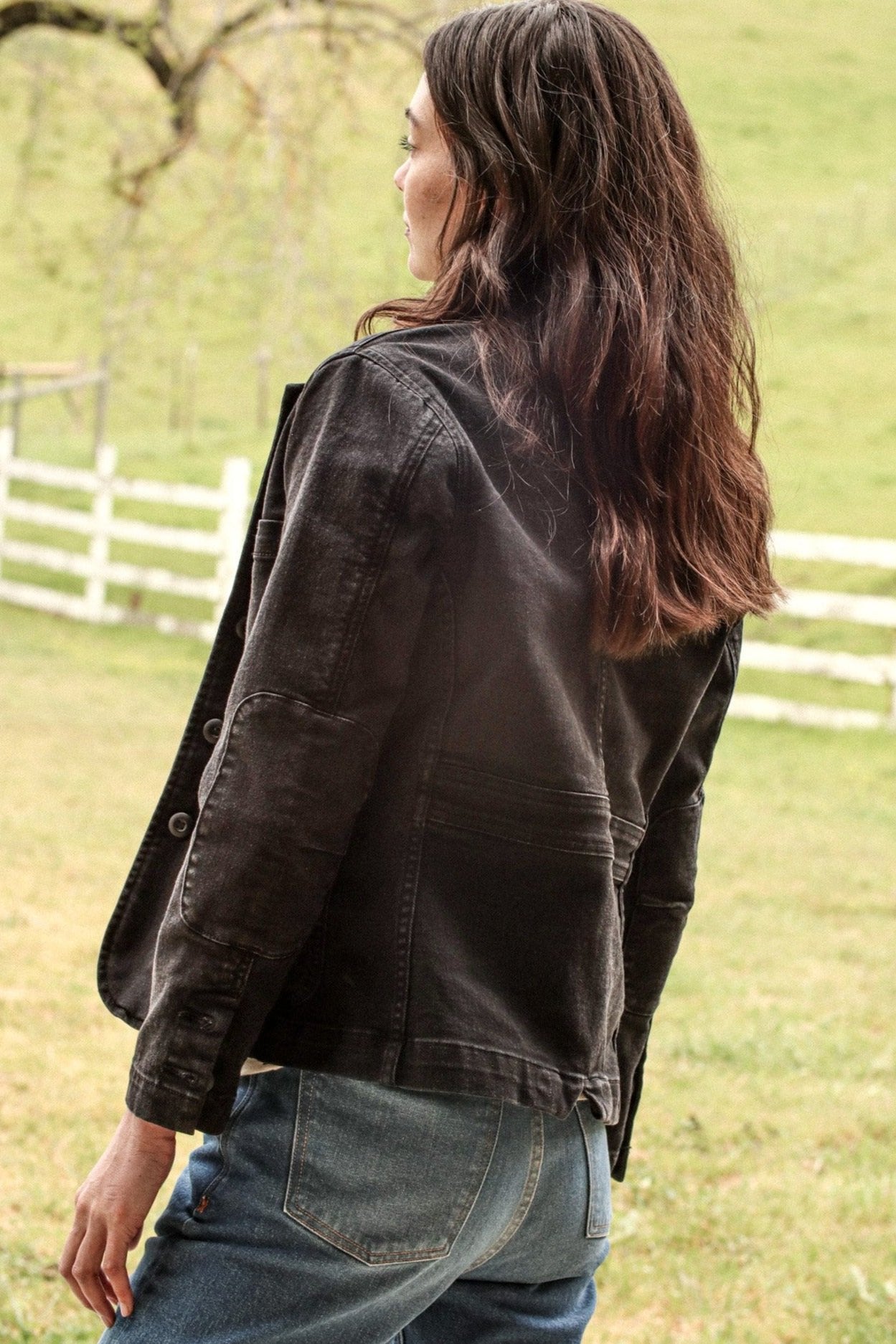 Back view of black denim chore blazer showing elbow patches and cuff buttons outdoors.