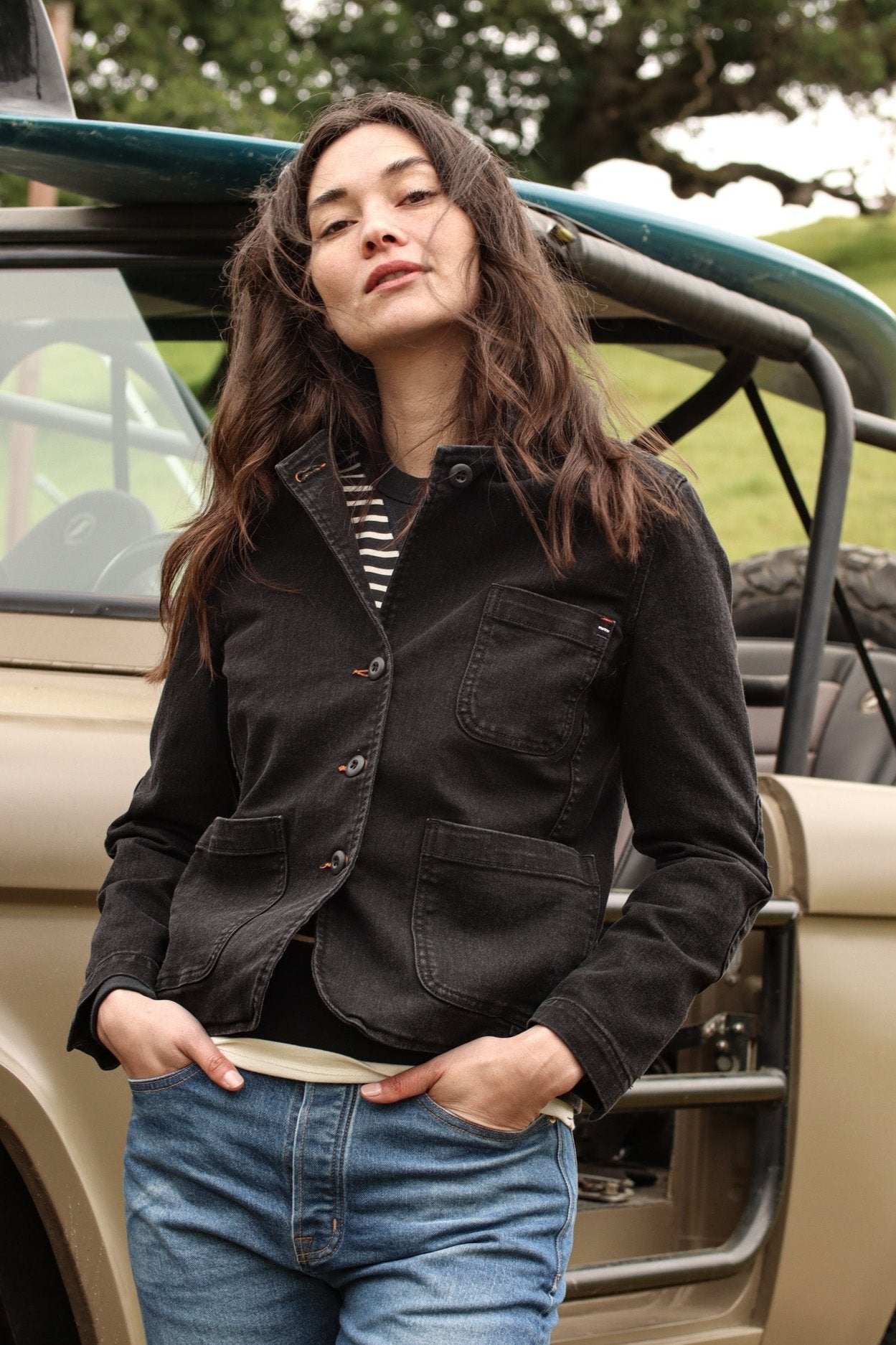 Woman in a black denim chore blazer and blue jeans leaning against an open-top vehicle with a surfboard.