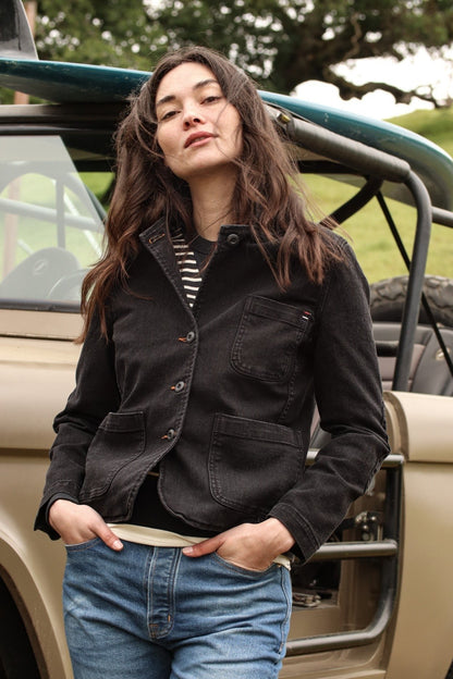 Woman in a black denim chore blazer and blue jeans leaning against an open-top vehicle with a surfboard.