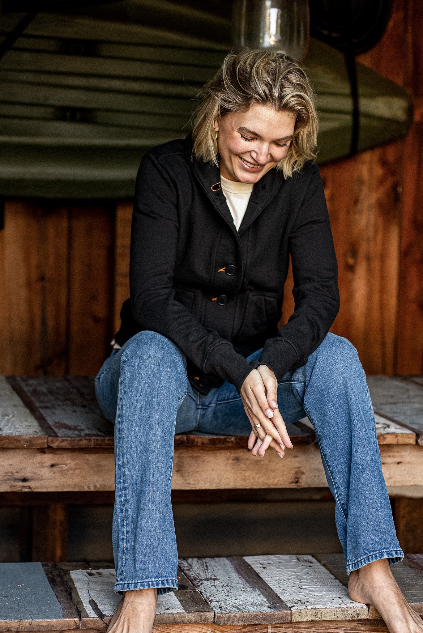 Woman in black Bodega French terry cardigan with orange contrast keyholes, paired with blue jeans on a rustic wood porch.