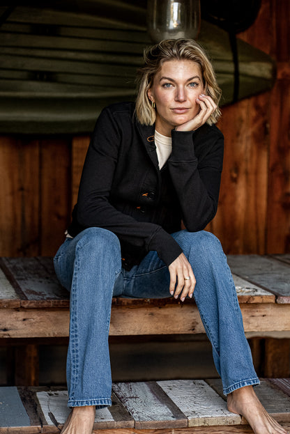 Woman in a black French terry Bodega cardigan with contrast tobacco stitching, paired with blue jeans, seated on a rustic.